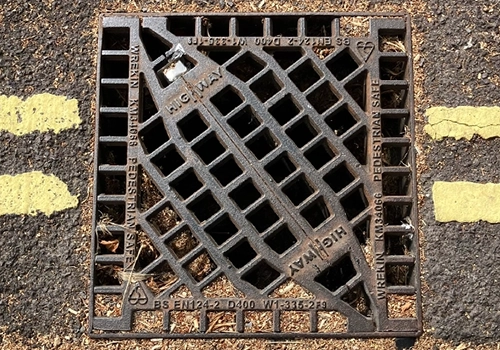 A square drainage gully on the road, with a square metal grate.