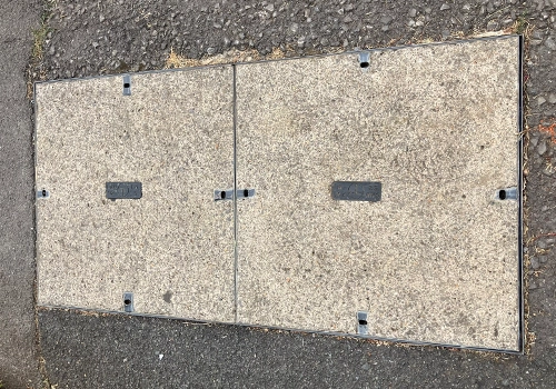 Rectangular telecoms cover, split into two concrete squares with a thin metal frame. A small rectangular plate in the centre of each square. 