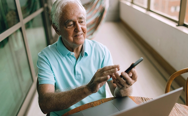 Elderly man sat at a table with a laptop while looking at his mobile phone