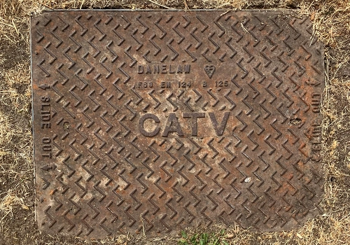 Rectangular CATV metal cover, within the ground, surrounded by dry grass. Featuring a raised tread pattern on the cover and the letters CATV.