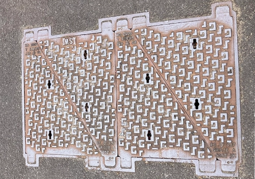 A large rectangular wastewater cover within the road surface. It has two metal panels, a raised geometric pattern and the words SW. 