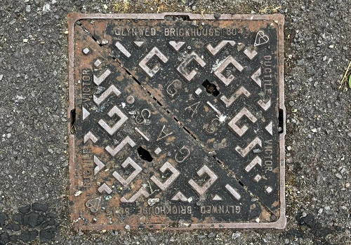 Square metal gas cover within the road. Featuring a raised geometric pattern and the word Gas within the centre. 